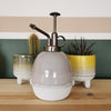 A gray plant mister with a hand finished texture, displayed on a wooden surface with two potted plants in the background.