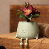 Cute green planter with a smiling face and a red flower on a wooden surface.